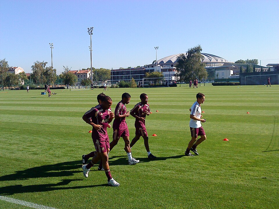 Four players in maroon training kit jogging on a grass pitch at Tola Vologe with floodlights behind them